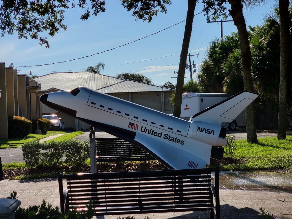 A scale model of a NASA space shuttle displayed outdoors in Cape Canaveral, Florida, surrounded by benches, palm trees, and buildings under a clear blue sky.