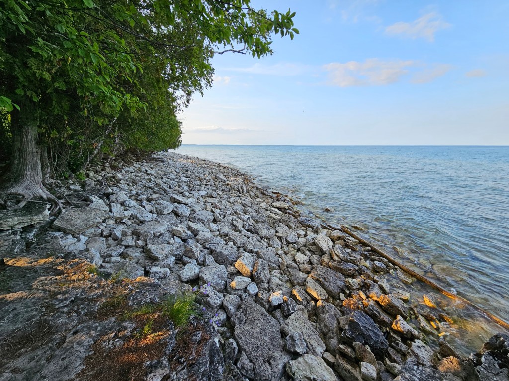 A tranquil shoreline at Cave Point, Wisconsin, along Lake Michigan, featuring rocky terrain, clear blue water, and lush green trees under a soft, sunny sky.