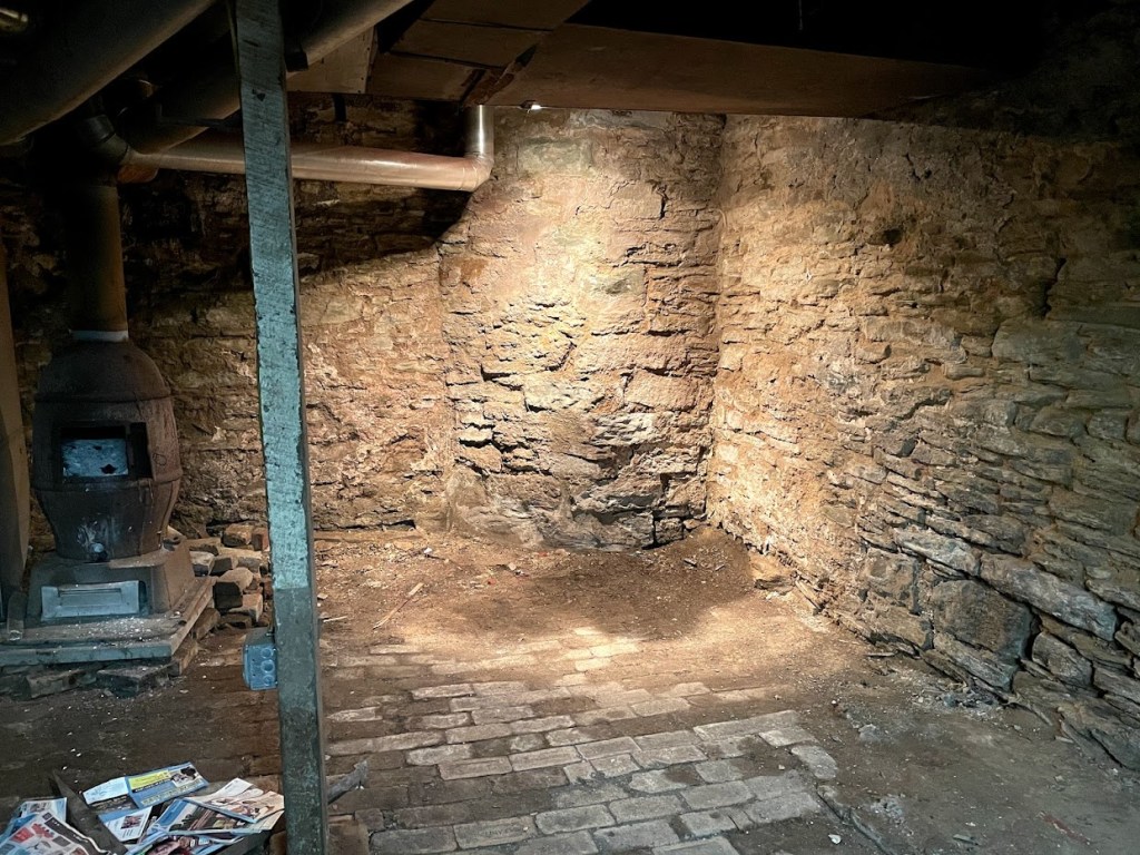 A basement in a 19th-century house in Perry North, featuring exposed stone walls, a vintage cast-iron stove, and a brick and dirt floor illuminated by soft, filtered light.