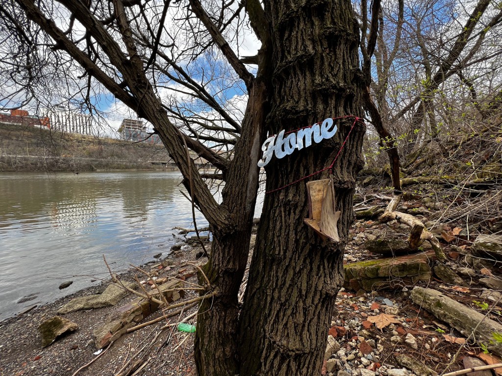 A tree along the Monongahela River in Pittsburgh, adorned with a decorative sign reading 'Home' and a broken branch that looks like an arrow pointing downward, surrounded by rocky and wooded terrain under a partly cloudy sky.