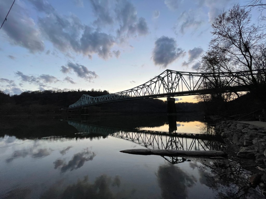 The Sewickley Bridge at dusk, viewed from the Sewickley side of the Ohio River, with its reflection mirrored on the calm water under a sky painted with soft clouds and fading light.