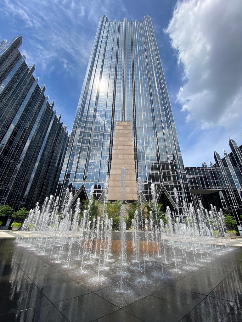 High-rise glass buildings at PPG Place in Pittsburgh reflect the sky and sunlight, with a vibrant fountain in the foreground spraying water in elegant patterns, under a mix of blue skies and fluffy clouds.