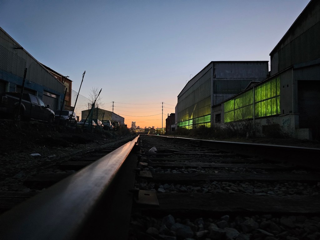 A low-angle view of railroad tracks at dusk, flanked by industrial buildings with glowing green windows on the right and silhouetted structures against a vibrant orange and blue sunset.