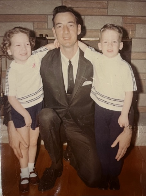 A vintage family photo featuring a father in a suit kneeling on the floor with his arms around two young children dressed in matching outfits, smiling in front of a stone fireplace.