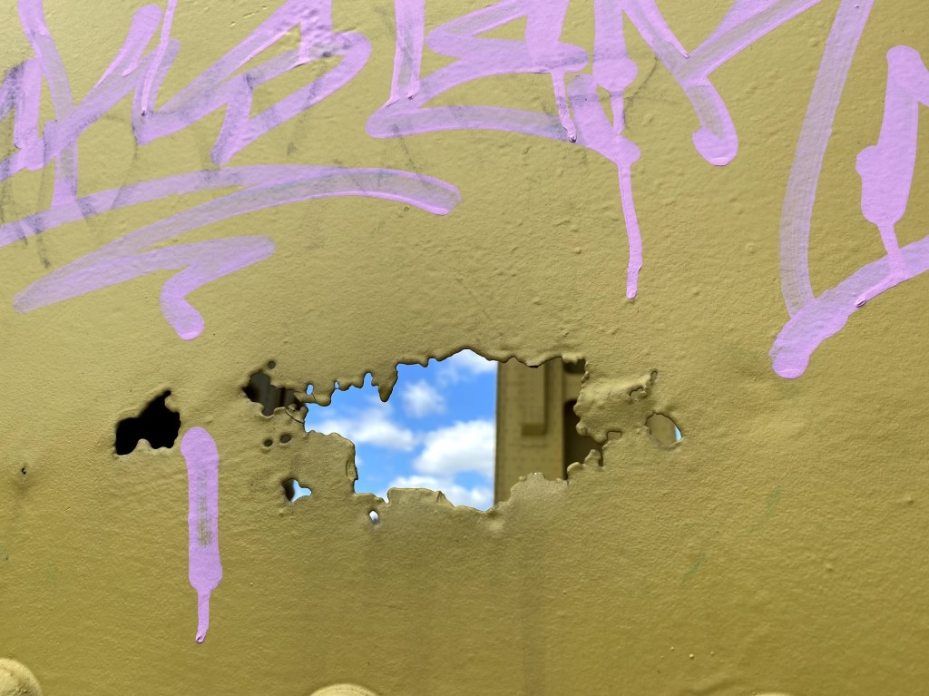 A close-up of a weathered, yellow-painted bridge with purple graffiti, featuring a jagged hole that reveals a glimpse of blue sky and clouds beyond.