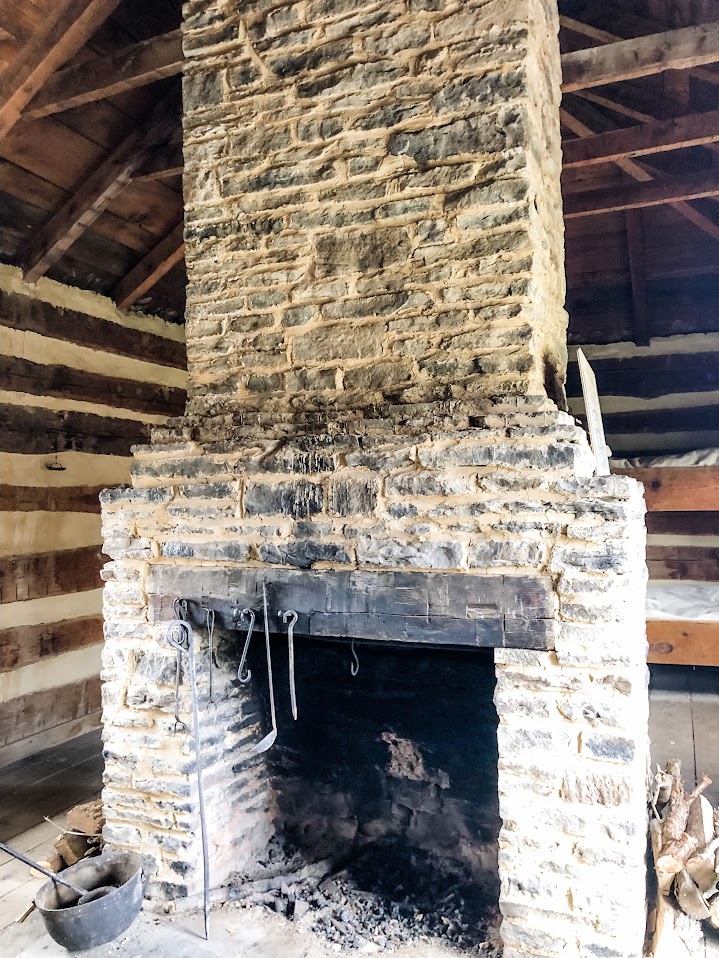 A rustic stone fireplace inside a historic log cabin, featuring an assortment of wrought iron tools and a blackened hearth, surrounded by exposed wooden beams and walls.