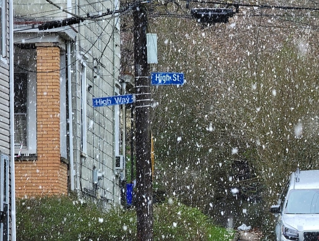 Snow falls gently on a street corner where 'High Way' and 'High St' signs intersect. The scene captures a quiet, wintry moment with houses and greenery lightly dusted by the snow.