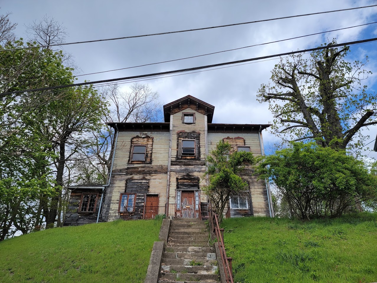A decaying three-story house sits atop a grassy hill, with a crumbling concrete staircase leading up to its rusted front doors. The house's wooden siding is faded and charred in places, with broken windows and missing panels exposing the structure beneath. Overgrown bushes partially obscure the facade, while trees with sparse leaves stand against a cloudy blue sky. Power lines stretch across the image, adding to the scene of abandonment and disrepair.