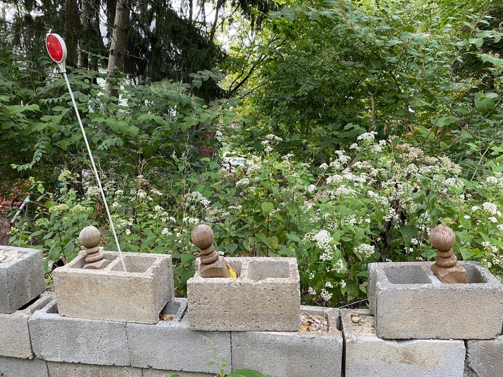 A makeshift barrier of cinder blocks with decorative wooden posts and a reflective marker, set against a backdrop of overgrown greenery and wildflowers.
