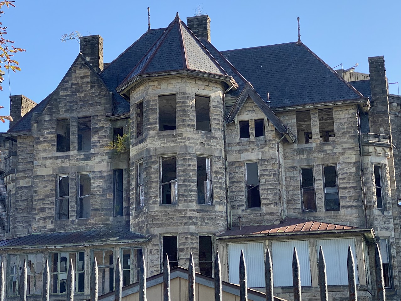 A massive, abandoned stone building with a gothic architectural style stands behind a spiked metal fence. The structure features a central turret, steeply pitched roofs, and multiple chimneys. Many windows are broken or missing, leaving dark, empty spaces in their frames. Vegetation grows from cracks in the stone walls and window ledges, adding to the eerie sense of decay. Rusted metal roofing and crumbling sections of the facade suggest years of neglect. The bright blue sky contrasts sharply with the haunting, weathered exterior of the building.
