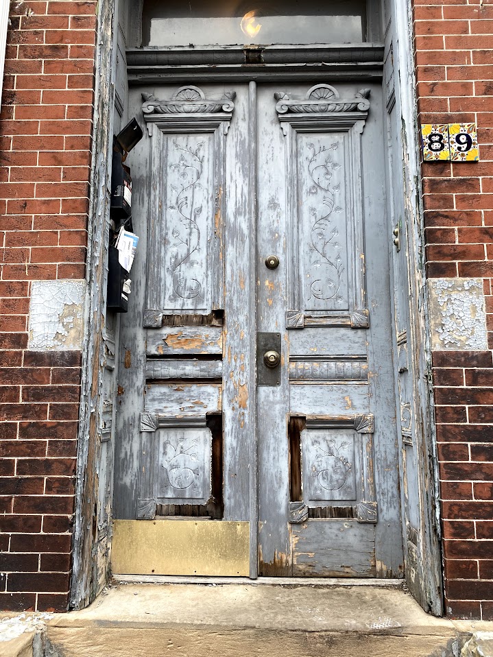 A set of weathered double doors with intricate carved floral and geometric details, faded blue-gray paint peeling to reveal the wood beneath. The bottom panels are damaged with missing sections. A brass kick plate is affixed to the lower part of the right door. Above, a transom window lets in dim light, reflecting a glow from inside. To the left, black metal mailboxes are mounted on the brick wall, overflowing with letters and papers. To the right, a colorful ceramic tile plaque displays the house number "89." The surrounding red brick facade has patches of peeling paint near the doorway.