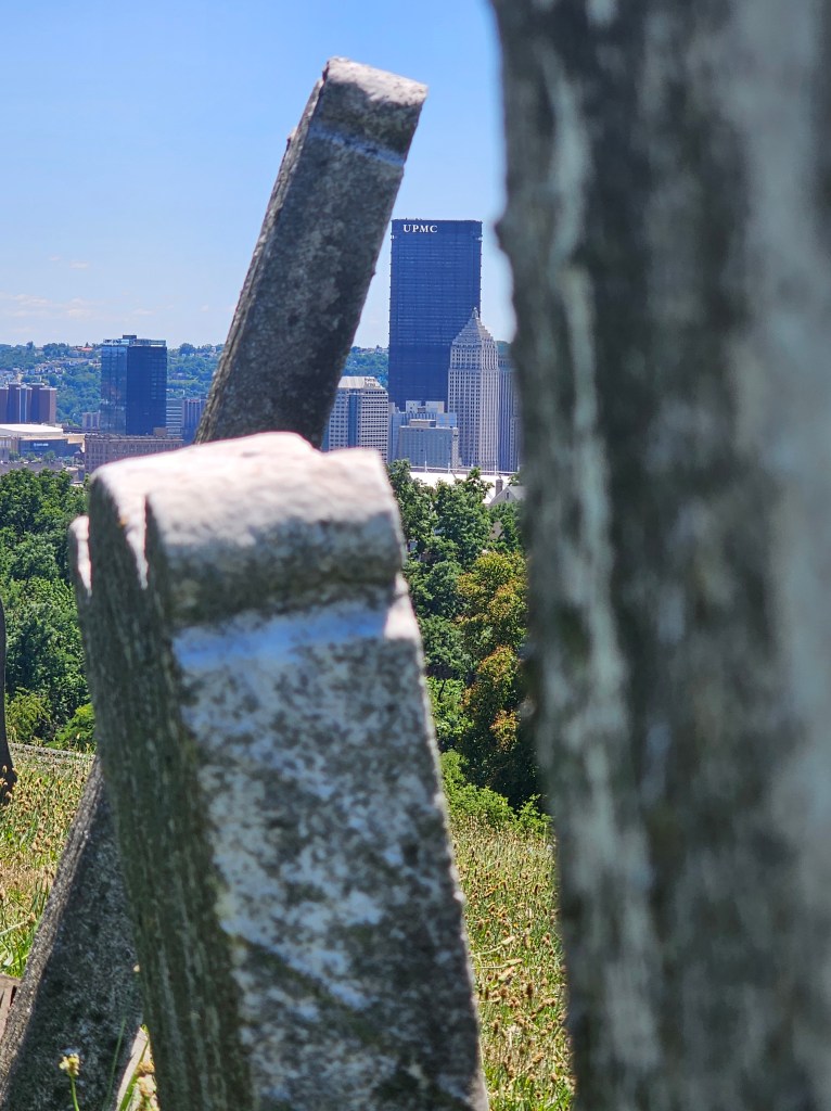 A weathered gravestone in the foreground leans diagonally, partially obscuring the view of Pittsburgh’s skyline in the background. The UPMC building and the Gulf Tower rise prominently behind the greenery of a summer hillside, creating a striking contrast between urban life and the stillness of the cemetery.