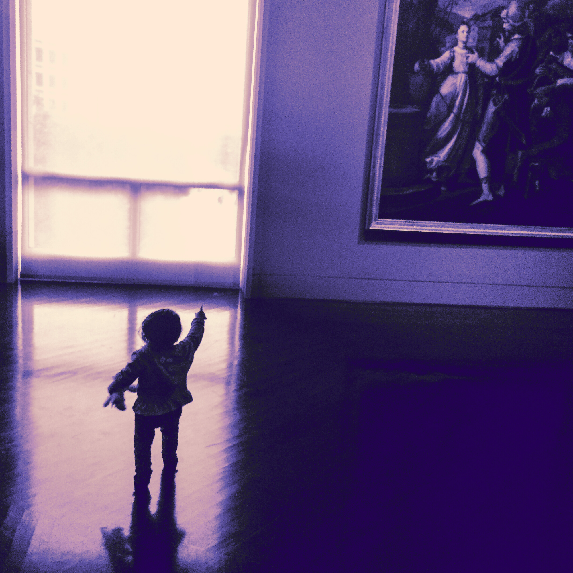 Silhouetted toddler pointing upward while standing in a gallery room with polished wooden floors. The child faces a large, glowing window, with a framed classical painting hanging on the wall to the right.
