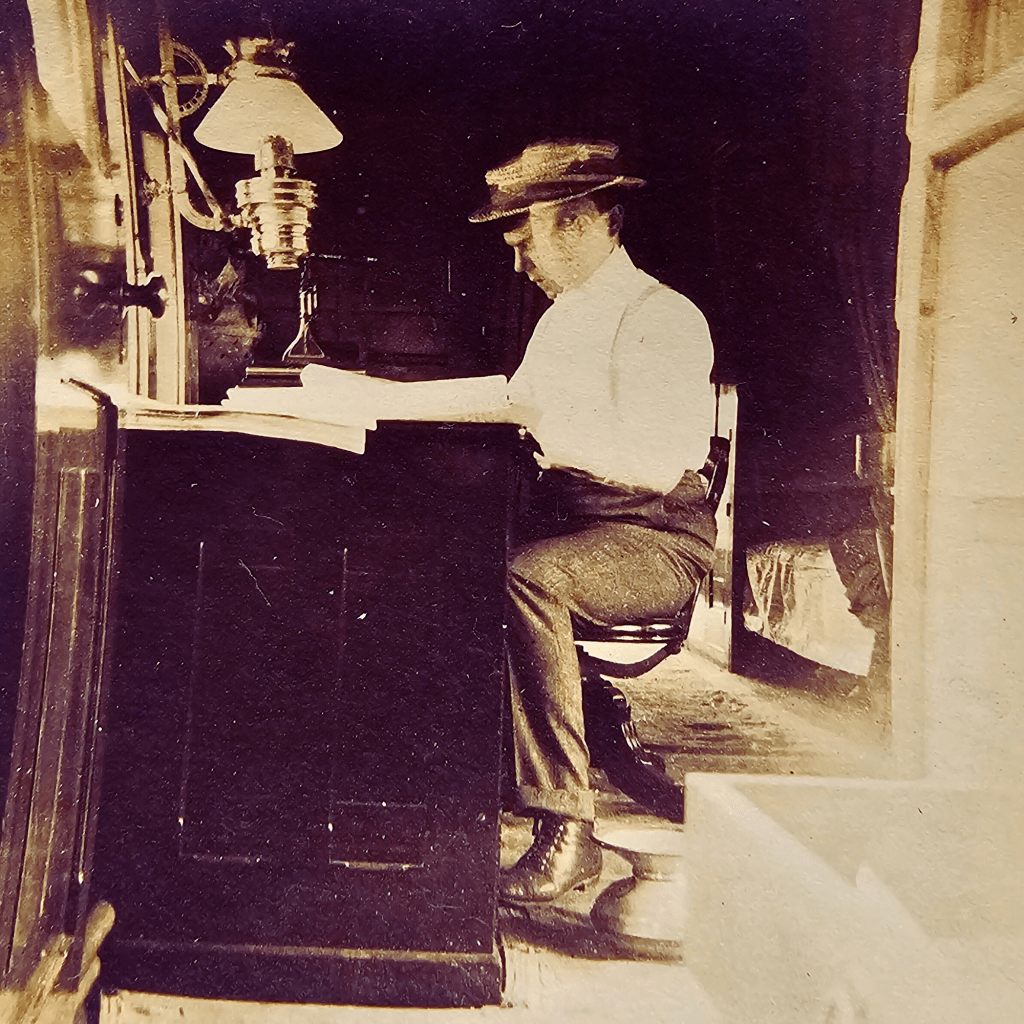 "Sepia-toned photograph of a man in early 20th-century attire sitting at a large wooden desk inside a dimly lit room or railcar. He is wearing a straw boater hat, white shirt, and dark trousers, focused intently on writing or reading a document. An ornate oil lamp sits on the desk, casting soft light over the workspace. The scene evokes a vintage, contemplative atmosphere."