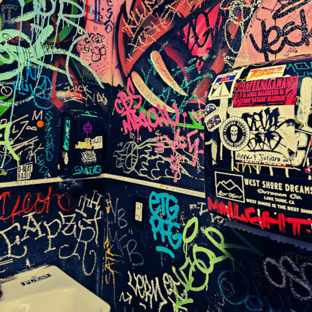 "A graffiti-covered public restroom wall filled with colorful spray paint tags, stickers, and street art. Layers of overlapping text in vibrant hues like pink, green, blue, and red create a chaotic, urban collage. Stickers advertising bands, radio stations, and skate shops cover a metal hand dryer or paper towel dispenser. The edge of a toilet tank is visible in the lower corner, grounding the setting in a gritty, underground aesthetic."