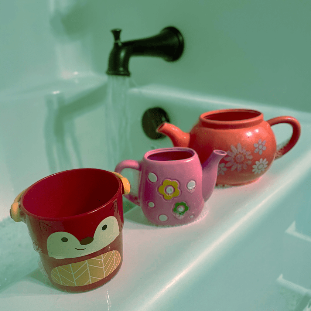 "Three colorful plastic toys—a red fox-faced bucket, a pink polka-dotted watering can, and an orange teapot with flower designs—sit lined up on the edge of a bathtub. Water flows from a dark bronze faucet in the background, with bubbles collecting around the toys, evoking a playful, childlike bath time scene."