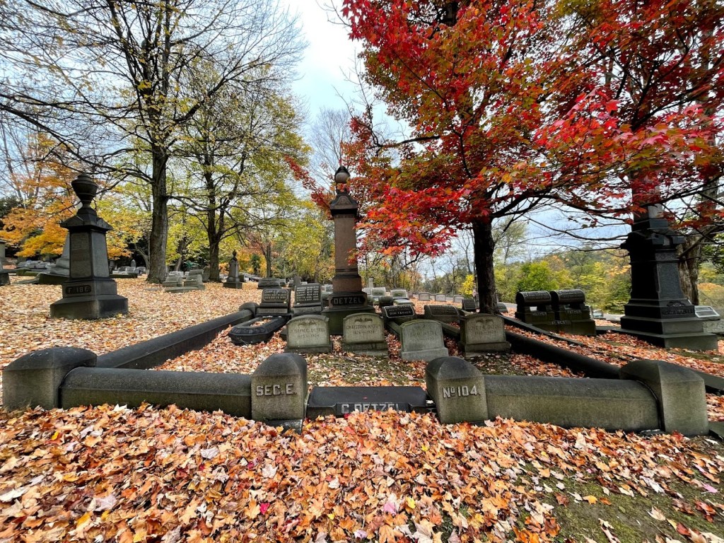 A quiet cemetery with a curved, rain-slicked brick path winding through rolling green hills. Tall trees with overhanging branches frame the image, while gravestones and monuments are scattered across the lush landscape. Misty skies and wet leaves add a somber, peaceful atmosphere.