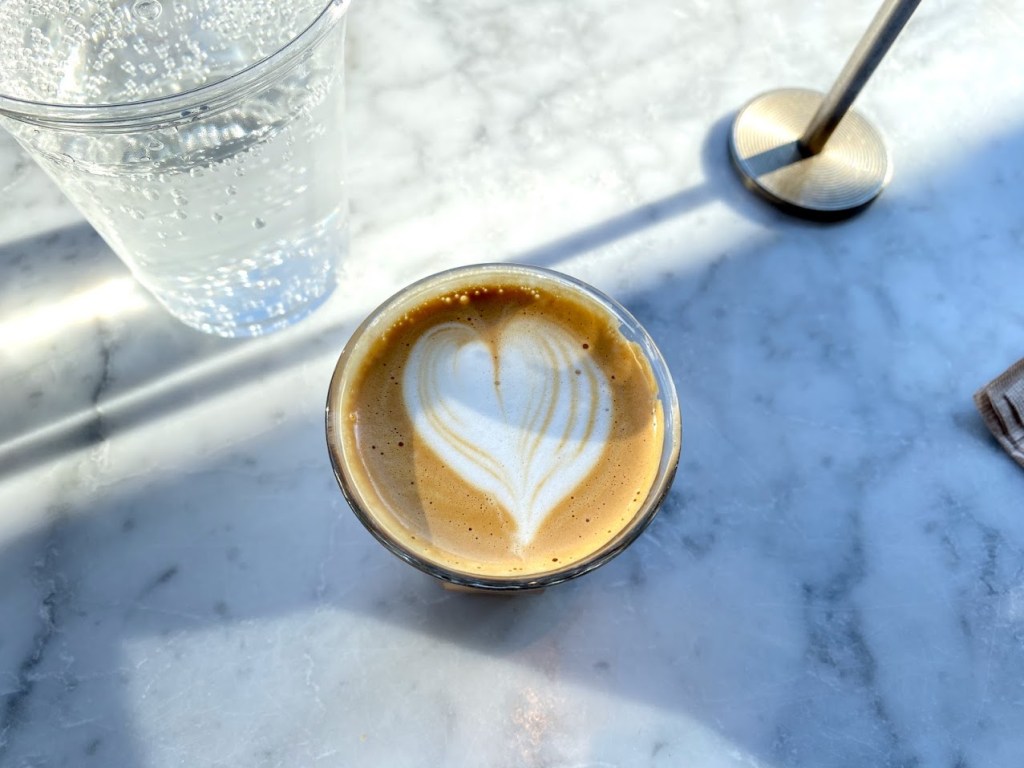 A small glass of espresso with heart-shaped latte art sits on a marble tabletop, bathed in soft natural light. To the left is a clear plastic cup of sparkling water, and a metal table lamp casts a shadow across the surface.