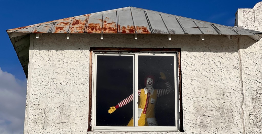 A weathered stucco building with a rusting metal roof and a large window shows a life-size Ronald McDonald figure inside, staring out with one arm raised in a wave. The clown's expression is frozen in a grin, creating an eerie contrast against the bright blue sky and cheerful pose.