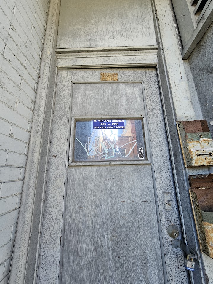 A weathered gray wooden door with a small rectangular window covered in graffiti and a sticker of a cartoon rabbit. Inside the window, a blue sticker reads, "ALL THE YEARS COMBINED 1965 to 1995 THEY MELT INTO A DREAM." The door is set in a cracked frame, surrounded by aged, peeling paint and adjacent to rusted metal fixtures, including a mailbox and a lockbox secured with a padlock. A white brick wall is visible to the left.