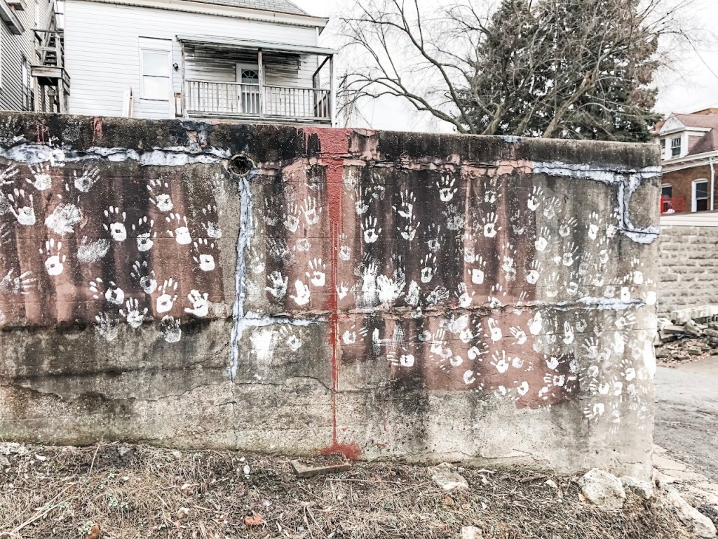 A weathered concrete retaining wall marked with numerous white handprints, some featuring painted red centers. A streak of red paint runs vertically down the center of the wall. Behind it, there is a white house with a small porch, leafless trees, and neighboring buildings. The scene has a somber, wintery atmosphere with muted colors and dry ground.