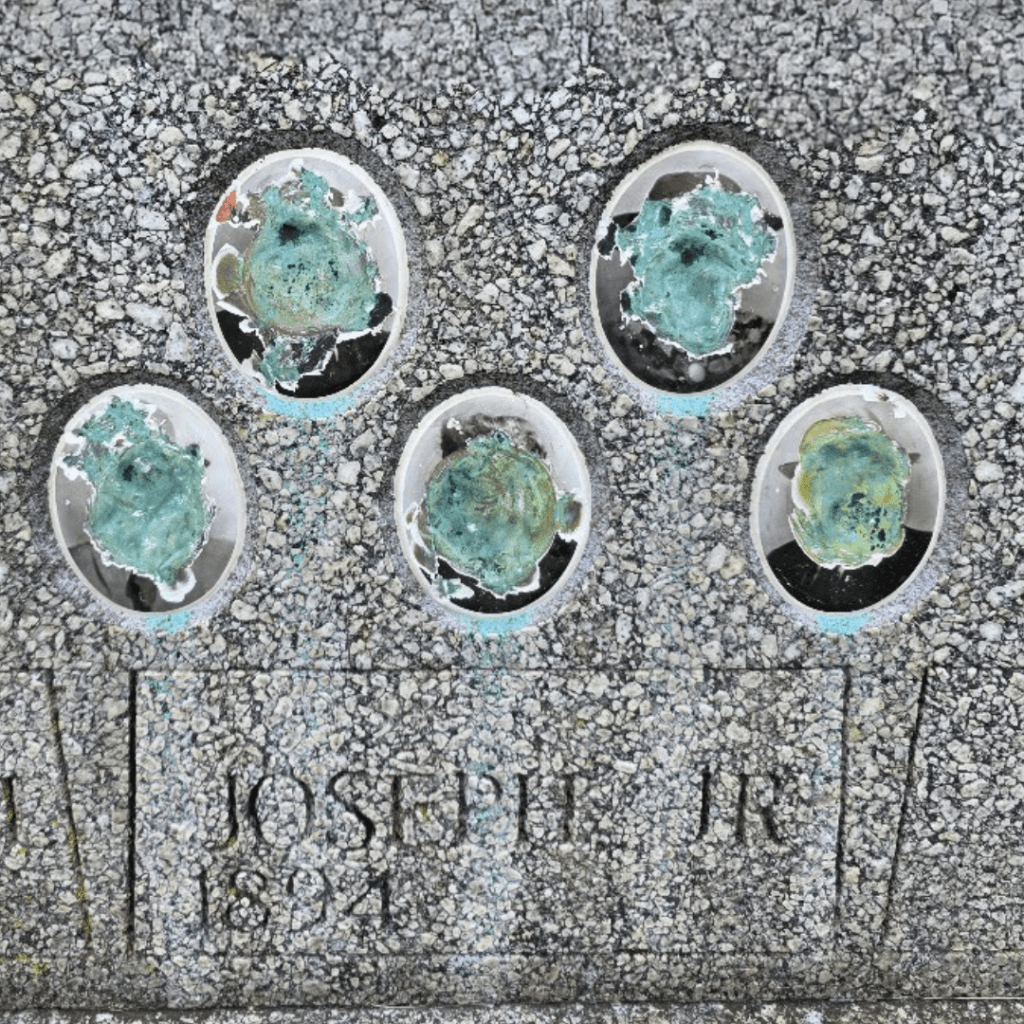 "Close-up of a weathered gravestone with five severely corroded oval photo frames, each obscuring what were likely ceramic portraits. Below the frames, the engraved name 'Joseph Jr' and the birth year '1894' are partially visible in a worn serif typeface on a speckled gray stone surface."