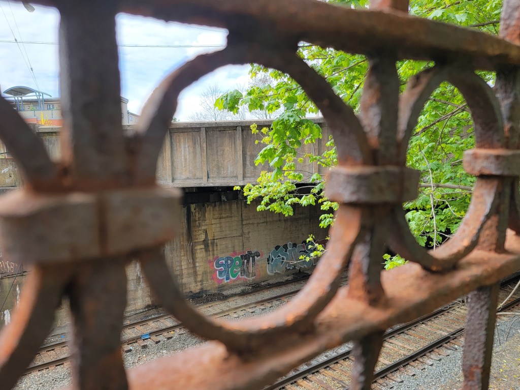 Urban scene of railroad tracks running below with train graffiti on a concrete wall beside the tracks, partially visible through rusty iron bars with circular patterns, surrounded by green foliage and trees.