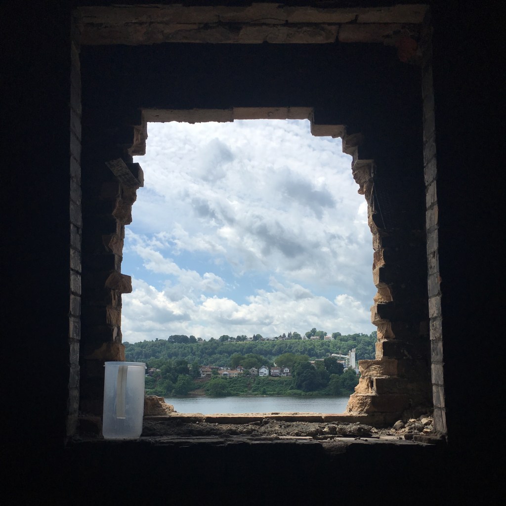 View through a jagged, partially demolished brick window framing a distant river, green hillside, and scattered houses under a sky filled with layered clouds. A translucent plastic pitcher sits on the rough ledge inside the dark room, adding contrast between decay and domesticity.