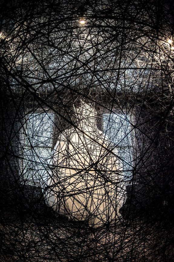 Backlit white figure seen through a dense web of black threads, like a cocooned silhouette inside an art installation.